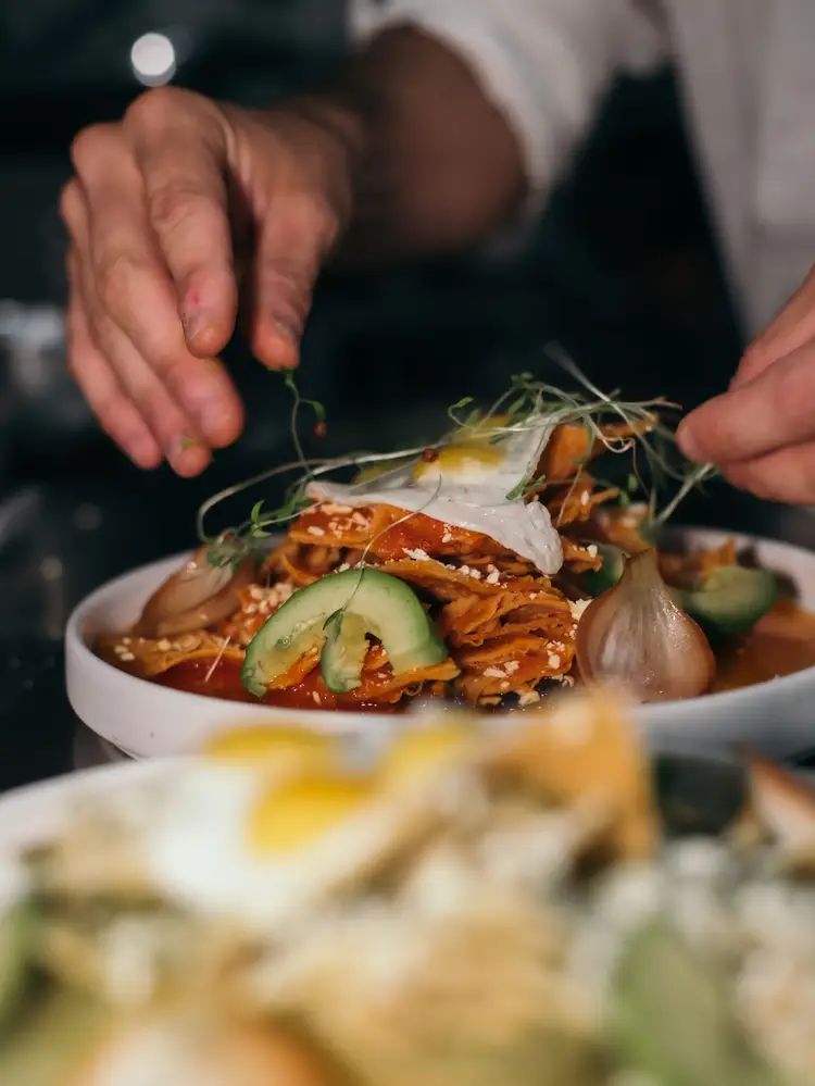 A chef garnishes a plated dish with shrimp and microgreens, layering toppings over sliced avocado and crispy tortilla