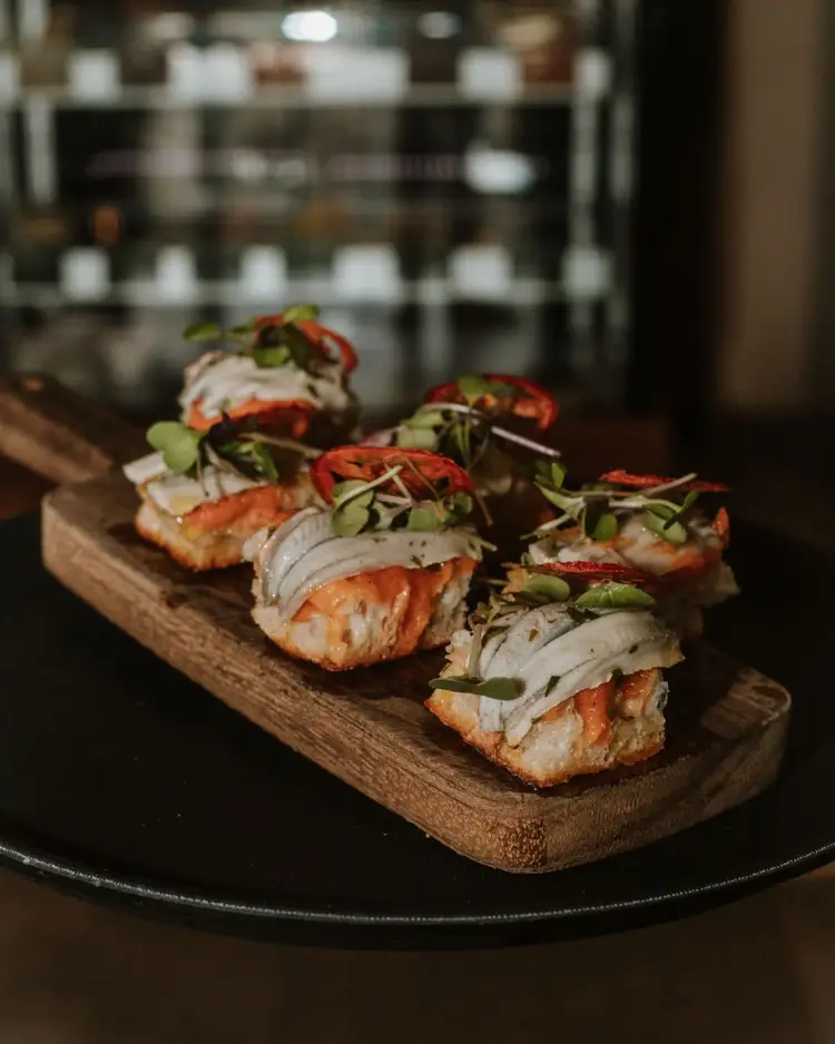 Four open-faced sandwiches with sliced meat, white cheese, red peppers and microgreens arranged on a wooden board atop a black plate in a restaurant kitchen
