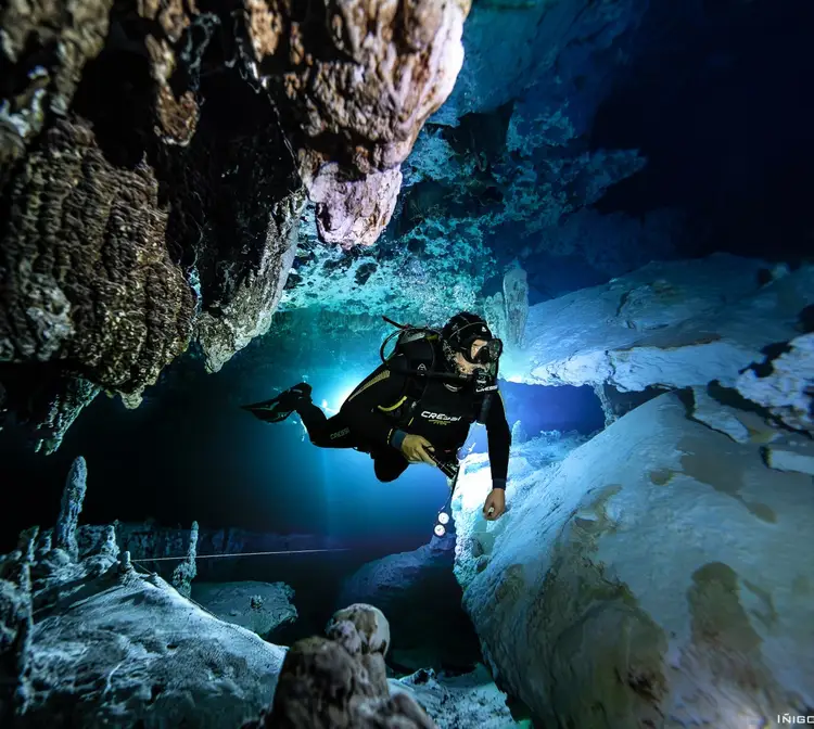 A scuba diver navigates through an underwater cave system with rocky limestone formations and turquoise water lit by dive lights A scuba diver navigates through an underwater cave system with rocky limestone formations and turquoise water lit by dive lights