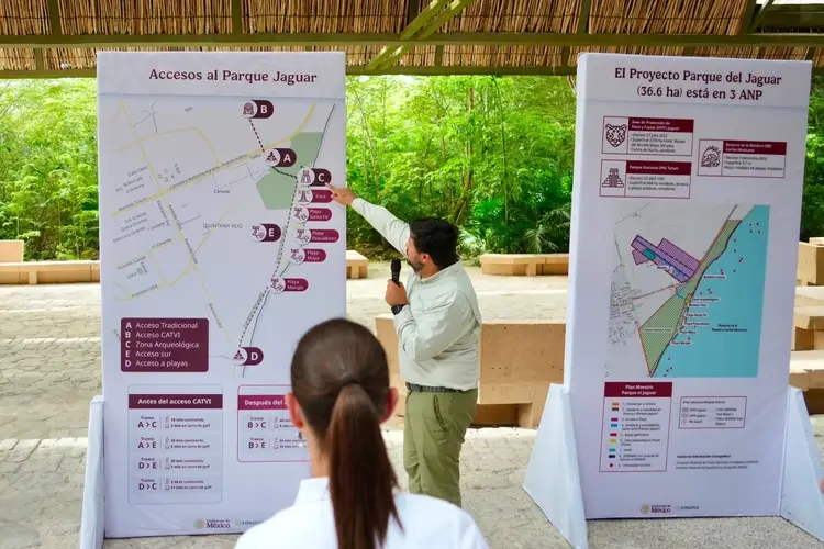 A man points to an informational sign about park access while a woman with long dark hair listens, standing beneath a thatched roof structure with forest visibl