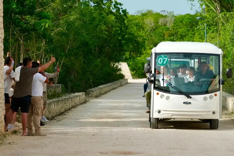 People stand beside a road waving at a white bus numbered 07 traveling through a tree-lined area