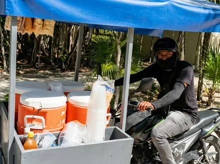 Person in black face covering and helmet sits on a motorcycle beside a street vending cart with orange coolers and cups under a blue tarp, with palm trees visib