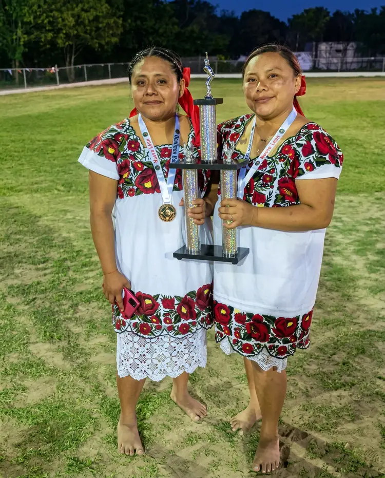 Two women wearing matching floral and white uniforms and medals hold a tall trophy while standing on a baseball field at dusk