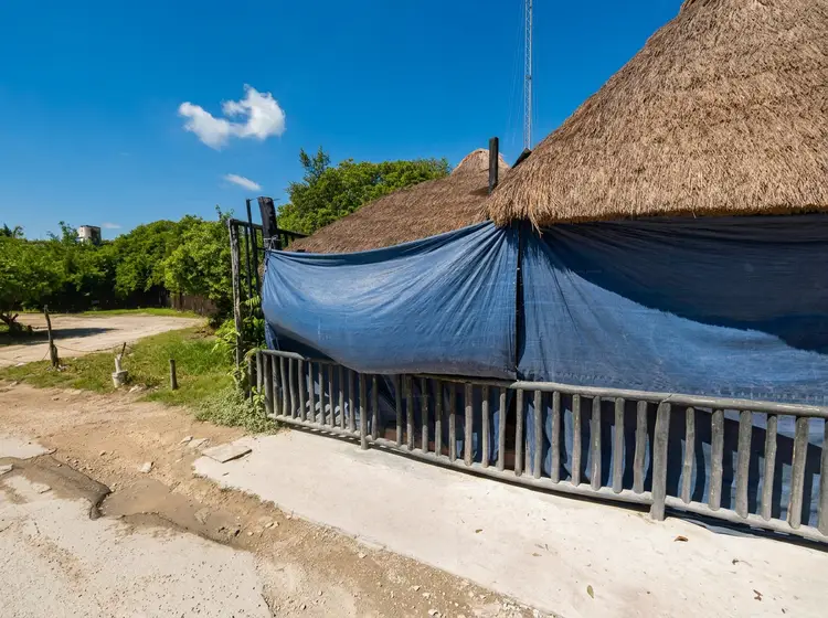 Blue tarp covers a thatched-roof beach structure behind a wooden fence in a sandy area with vegetation and clear sky