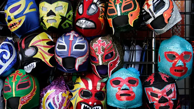 Colorful wrestling masks with various designs displayed on a dark wall, including blue, red, green, yellow and turquoise masks with bold patterns and eye holes