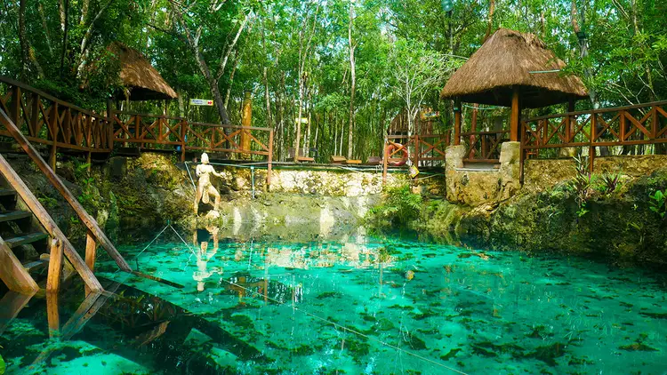 A clear turquoise cenote surrounded by wooden structures with thatched roofs, wooden railings, and lush green forest canopy A clear turquoise cenote surrounded by wooden structures with thatched roofs, wooden railings, and lush green forest canopy
