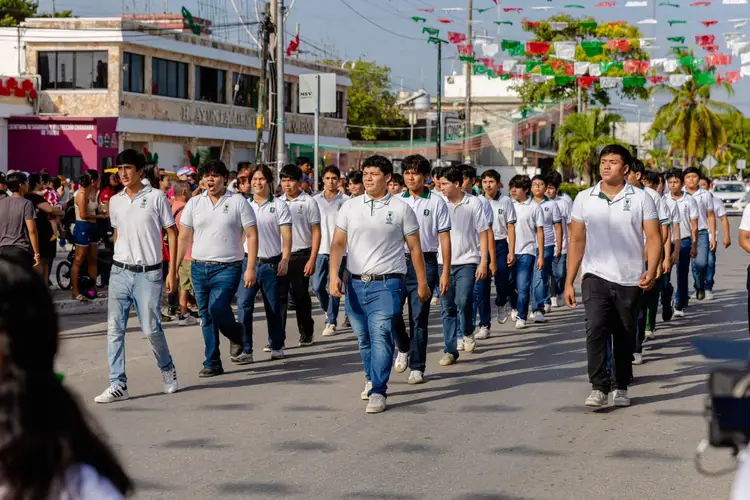 Large group of students in white polo shirts and blue jeans marching down a city street during a daytime parade with buildings and decorative flowers visible Large group of students in white polo shirts and blue jeans marching down a city street during a daytime parade with buildings and decorative flowers visible