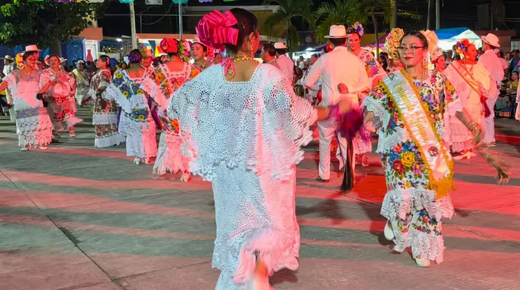 Grupo numeroso de personas con elaborados trajes de encaje rosa y colores vivos, y sombreros, baila sobre una alfombra roja en una plaza al aire libre Grupo numeroso de personas con elaborados trajes de encaje rosa y colores vivos, y sombreros, baila sobre una alfombra roja en una plaza al aire libre