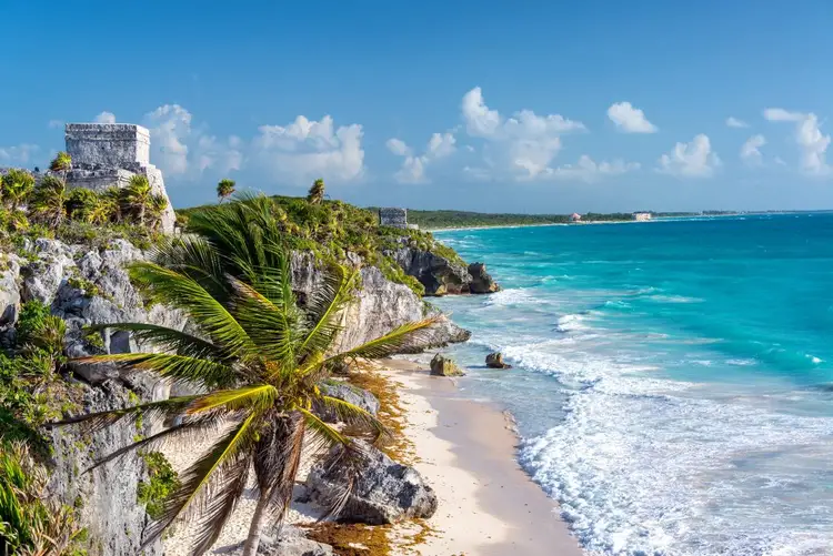 White stone ruins sit atop a cliff overlooking a sandy beach and turquoise water, with palm trees and tropical vegetation on the rocky coastline