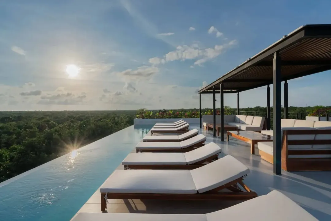 Tulum’s Real Estate Market Navigates Vacation Rental Over-Supply Challenges Lounge chairs line an infinity pool overlooking a forested landscape, with a covered seating area and pergola on a modern rooftop terrace