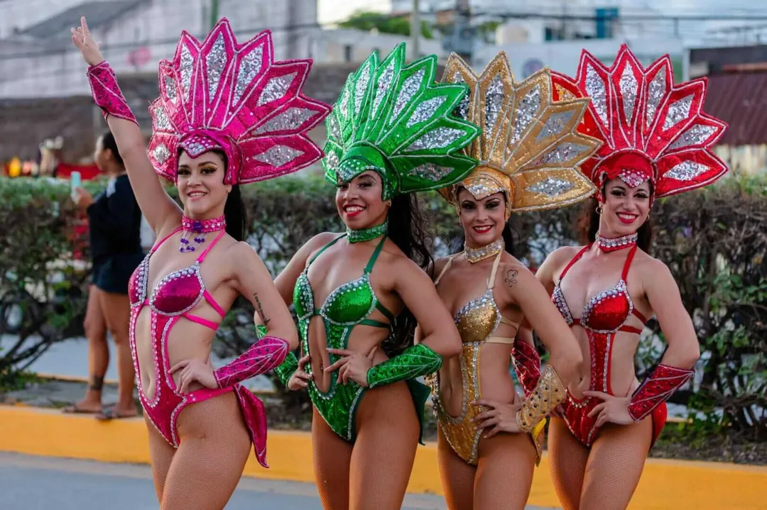 Four performers in sequined bikinis and elaborate feathered headdresses in pink, green, gold, and red stand posing outdoors at a carnival celebration