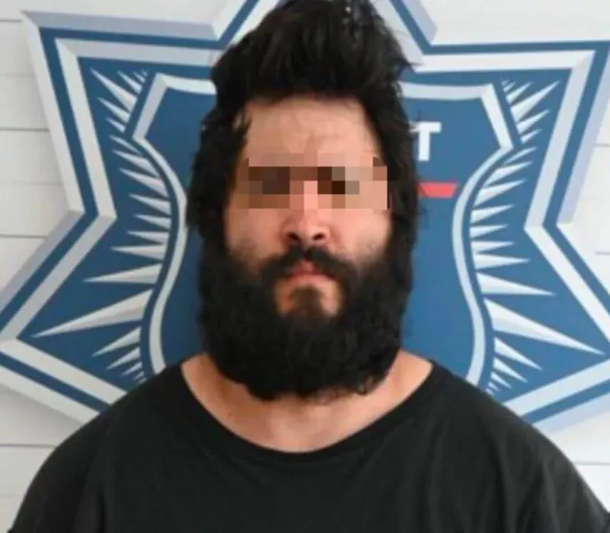 Foreign Perspectives Shape Growing Communities in Yucatan Peninsula Man with long black hair and beard wearing black shirt, face pixelated, posed in front of blue and white police or official badge logo