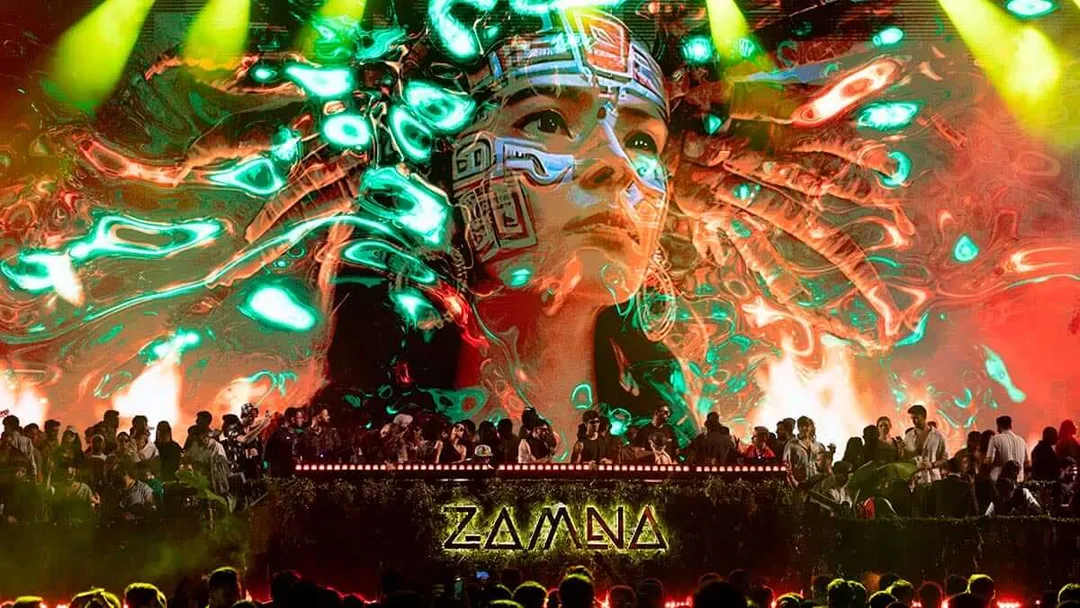 Large illuminated stage display showing a face with geometric patterns and neon green and red lighting effects, with festival attendees silhouetted in the foreg