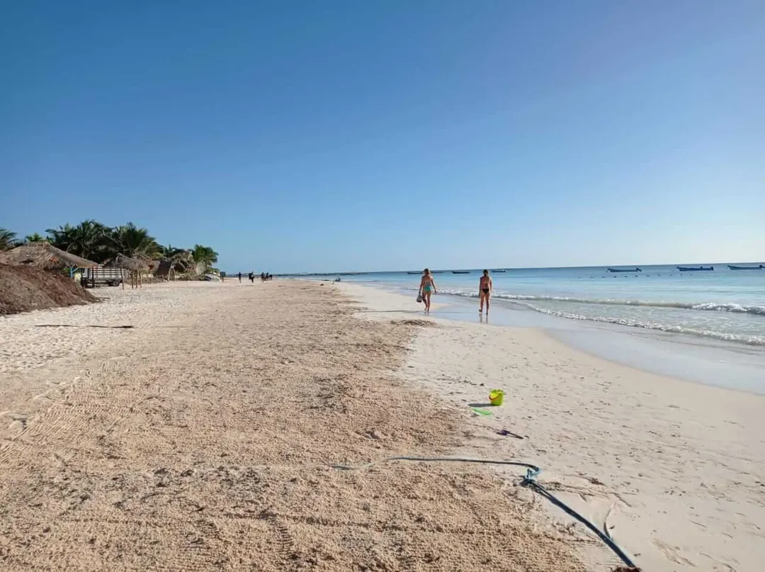 Beach with scattered seaweed and a yellow ball in the foreground, while two people wade in shallow water and boats sit offshore under clear sky
