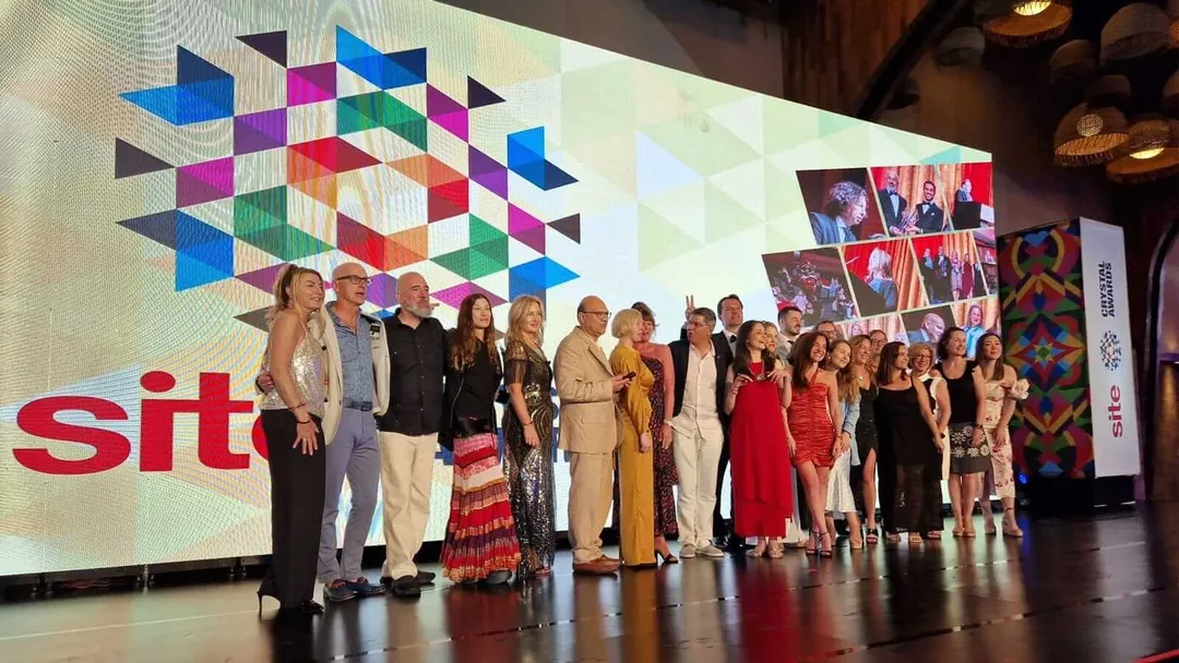 Incentive Travel Innovators Gather for Landmark 2025 Summit A group of people stand on stage in front of a large screen displaying a colorful geometric logo and partial text, with photographs and decorative panels visibl