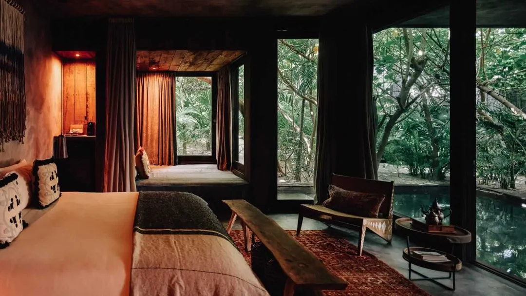 A modern bedroom with large windows overlooking trees features a bed with orange bedding, dark curtains, and minimalist furniture arranged on a red-toned rug