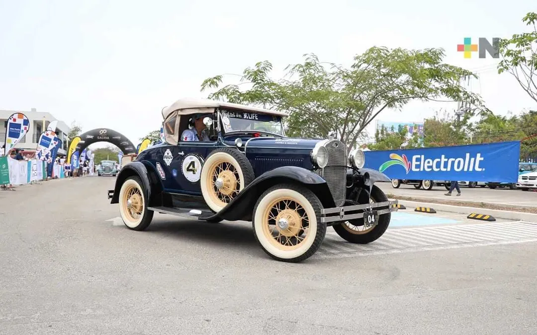A vintage dark blue car with the number 4 on its side drives through a rally checkpoint with spectators and sponsor banners visible along the roadside