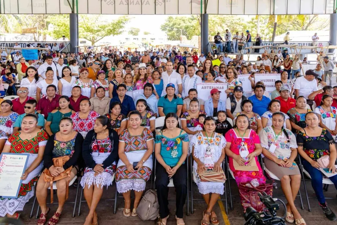 Large group of people gathered under a covered outdoor structure, with seated attendees in front wearing colorful traditional clothing and standing attendees fi