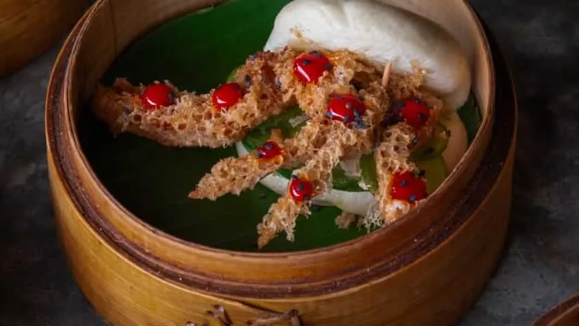 Fried shrimp pastries topped with red chili peppers sit on a green leaf inside a wooden steamer basket with a white cloth