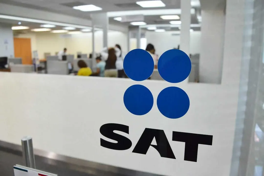 Free Tax Guidance Empowers Tulum Entrepreneurs Company logo with four blue circles above black letters on glass partition in an office with employees at desks in background