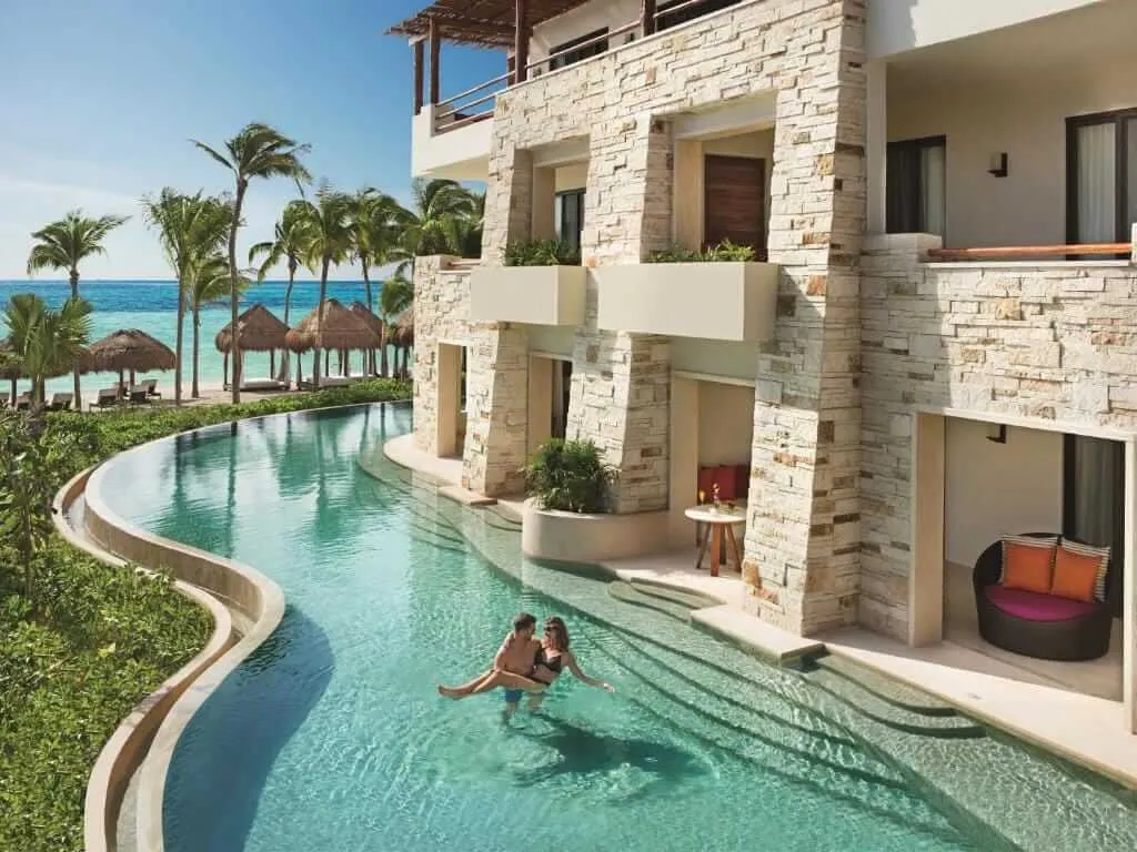 Tulum All-Inclusive Resorts Two people swim in a curved pool beside a multi-story stone building with ocean and palm trees visible in the background