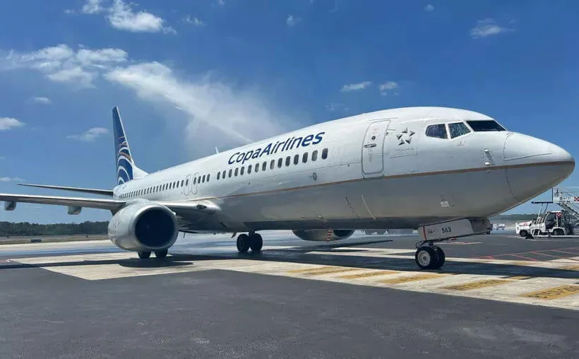 White commercial airliner parked on an airport tarmac under clear blue sky with service vehicles nearby