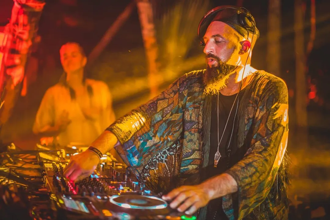Day Zero Festival Set for January 10, 2026, in Tulum DJ wearing headphones and patterned jacket operates equipment at a nightclub event with warm stage lighting and other people visible in the background