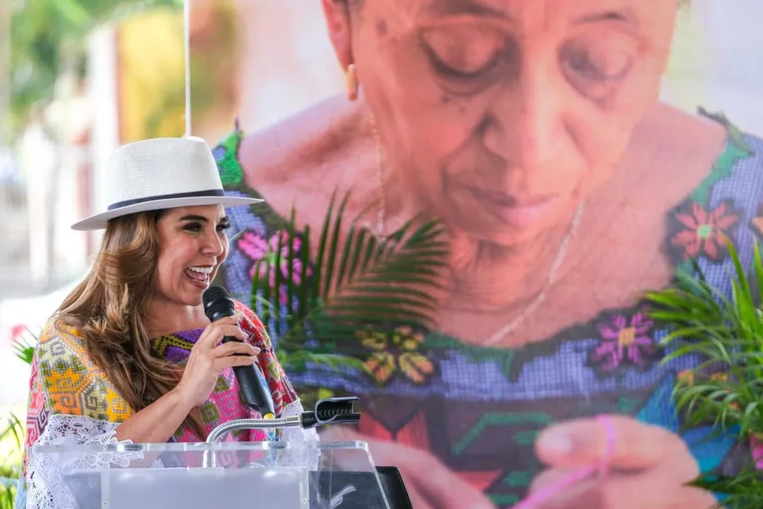 Tulum Honors Handicraft Masters Amidst Coastal Breeze Woman wearing white hat speaking into microphone at podium, with large projected image of person behind her