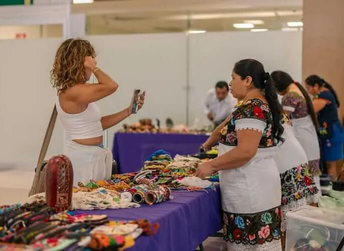 Tulum Airport Showcases Local Artisans’ Craftsmanship A woman in a white top examines colorful handmade textiles and crafts displayed on a purple table at an indoor market or fair