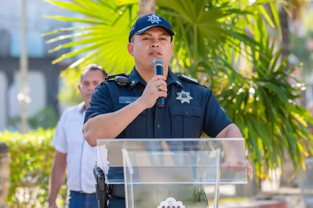 Tulum Hoteliers Condemn Rising Violence Amid Officer’s Death Law enforcement officer in uniform speaks at a podium with microphone outdoors, with palm trees and another person visible in the background