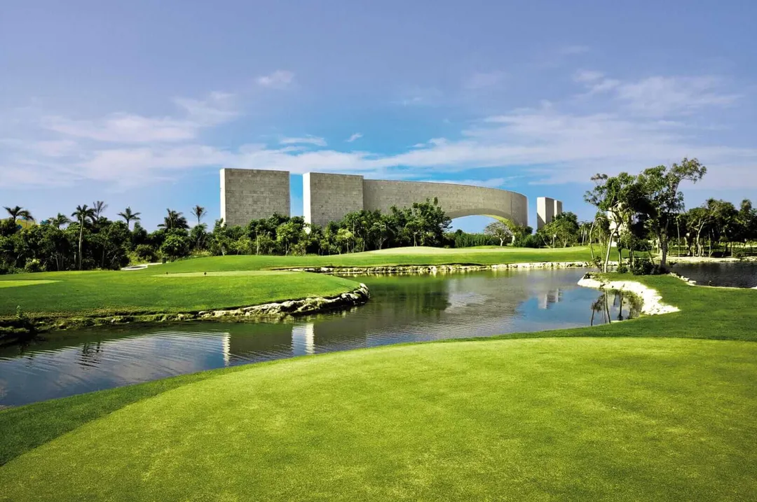 Modern concrete building with curved architecture sits across a manicured golf course with a water hazard and palm trees under blue sky