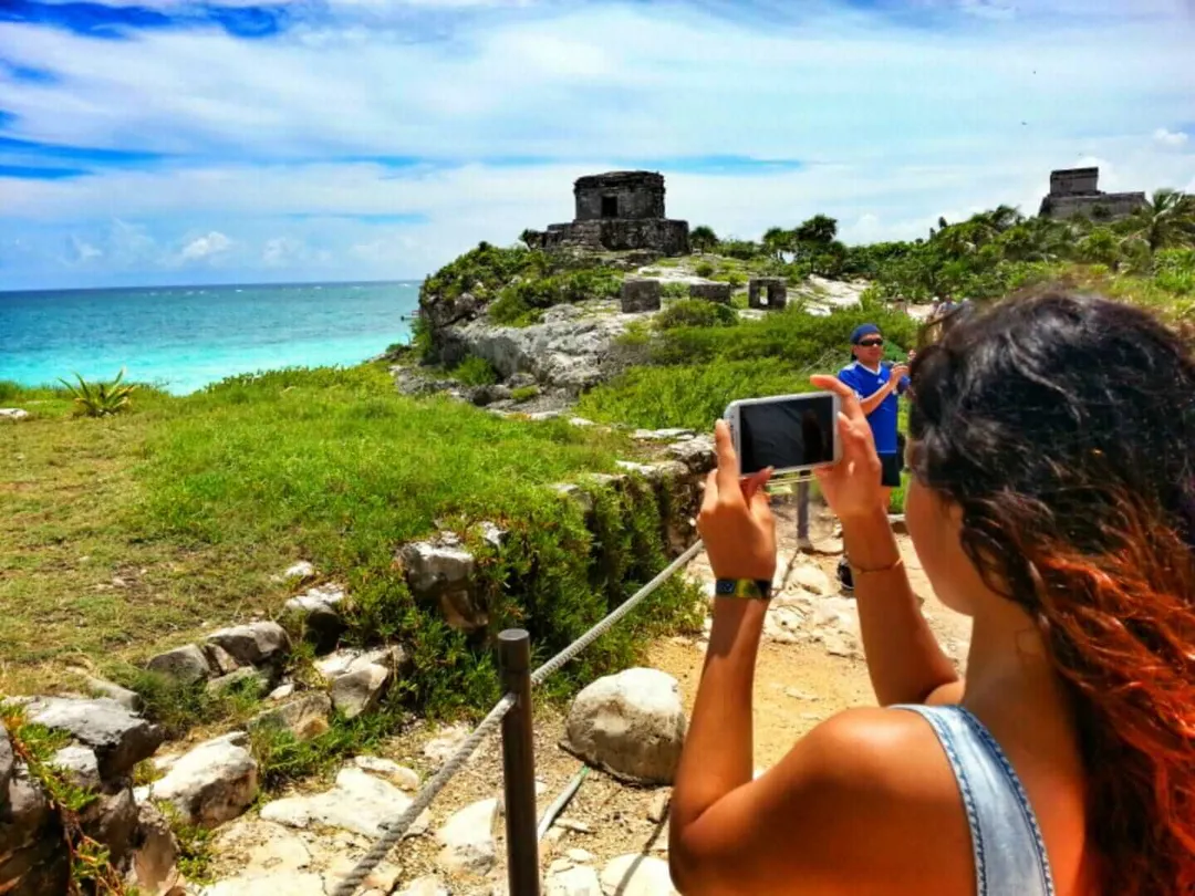 A visitor photographs ruins on a clifftop overlooking turquoise water and tropical vegetation