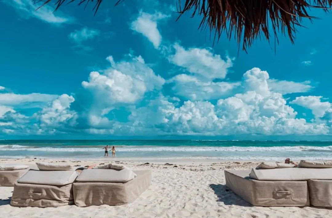 Two people stand in shallow ocean water while beach lounges with cushions sit in the sand under a thatched roof structure, with white clouds dotting a blue sky