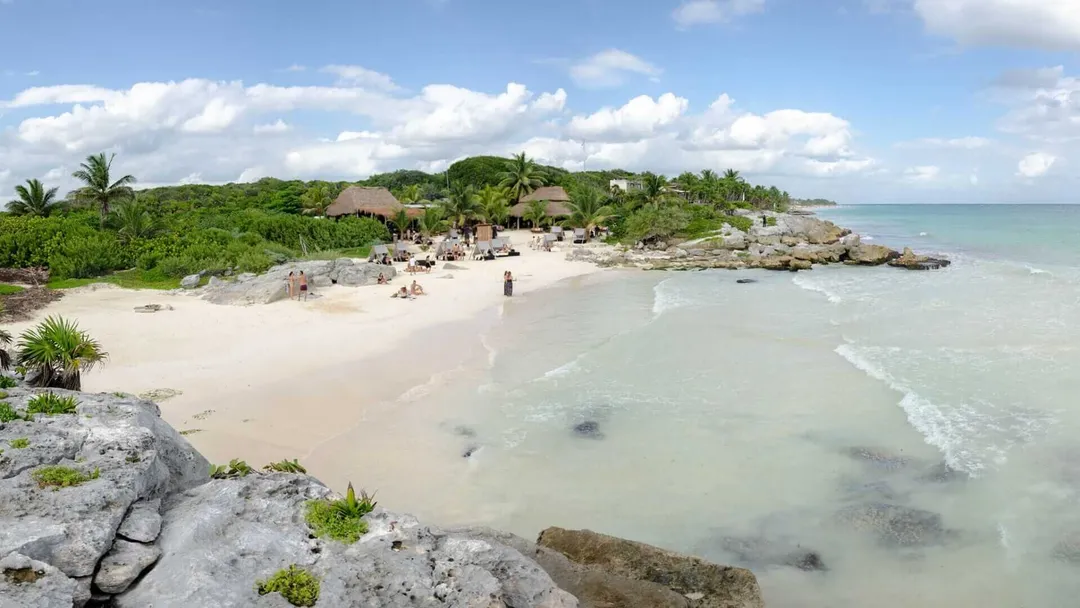 Beach with thatched-roof structures and visitors along the shoreline, surrounded by tropical vegetation and rocky outcrops