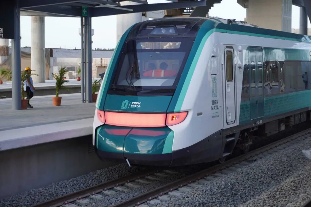 A teal and white commuter train stopped at a platform station with a person and potted plant nearby