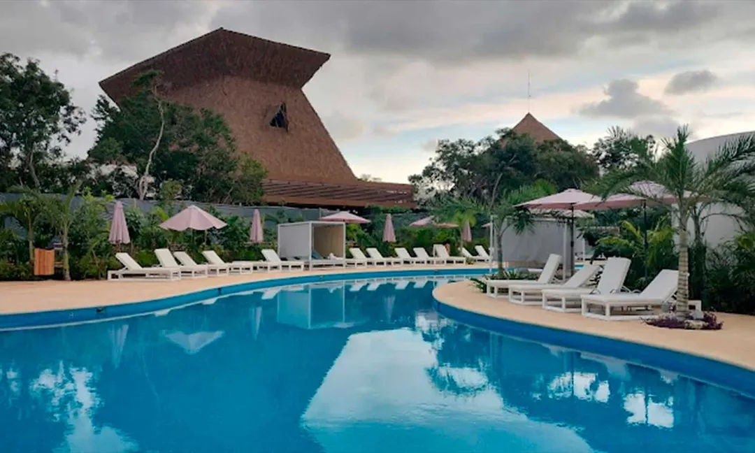 Maya Train Hotels Aim to Outshine Yucatan’s Luxury Resorts A large swimming pool reflects the sky in front of a distinctive thatched-roof building, with white lounge chairs and umbrellas lining the deck