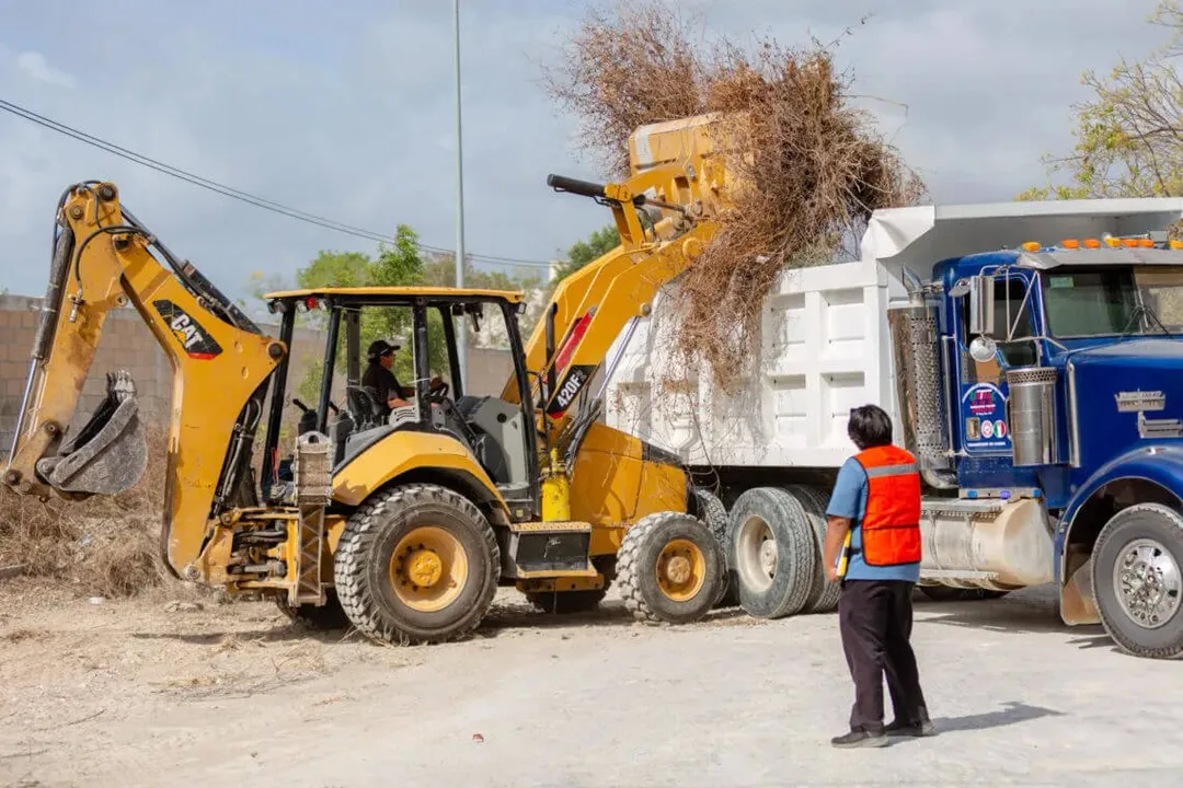 Yellow excavator loads debris into a dump truck while a worker in a safety vest supervises at a construction or cleanup site