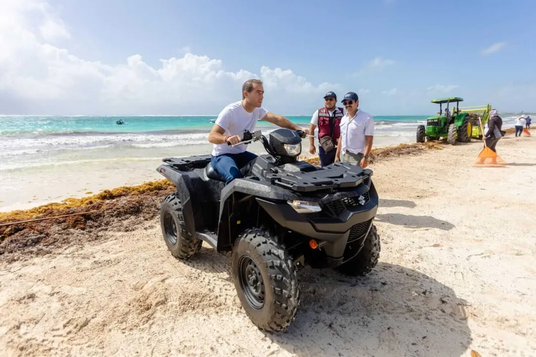 Man operates an all-terrain vehicle on a beach next to sargassum seaweed, with cleanup workers and equipment visible along the shoreline