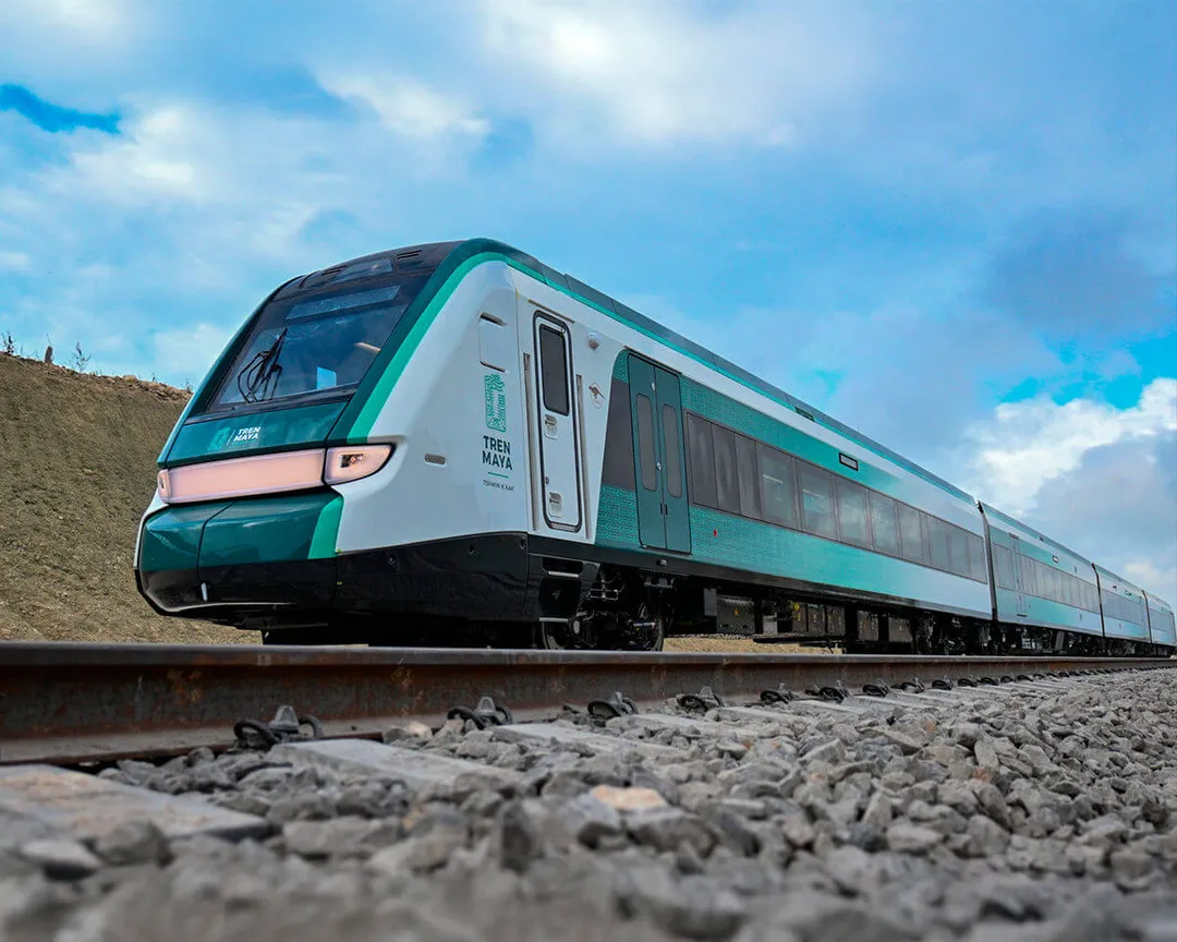 Tren Maya Launches Easter Holiday Travel Packages to Promote Regional Exploration A teal and white passenger train travels along railroad tracks under a blue sky with scattered clouds
