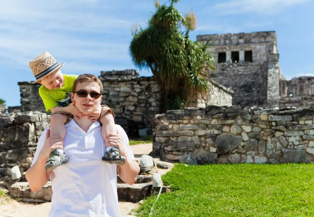 Tulum Braces for Holiday Traveler Wave An adult wearing sunglasses carries a young child on their shoulders while standing in front of ancient stone ruins and grassy grounds
