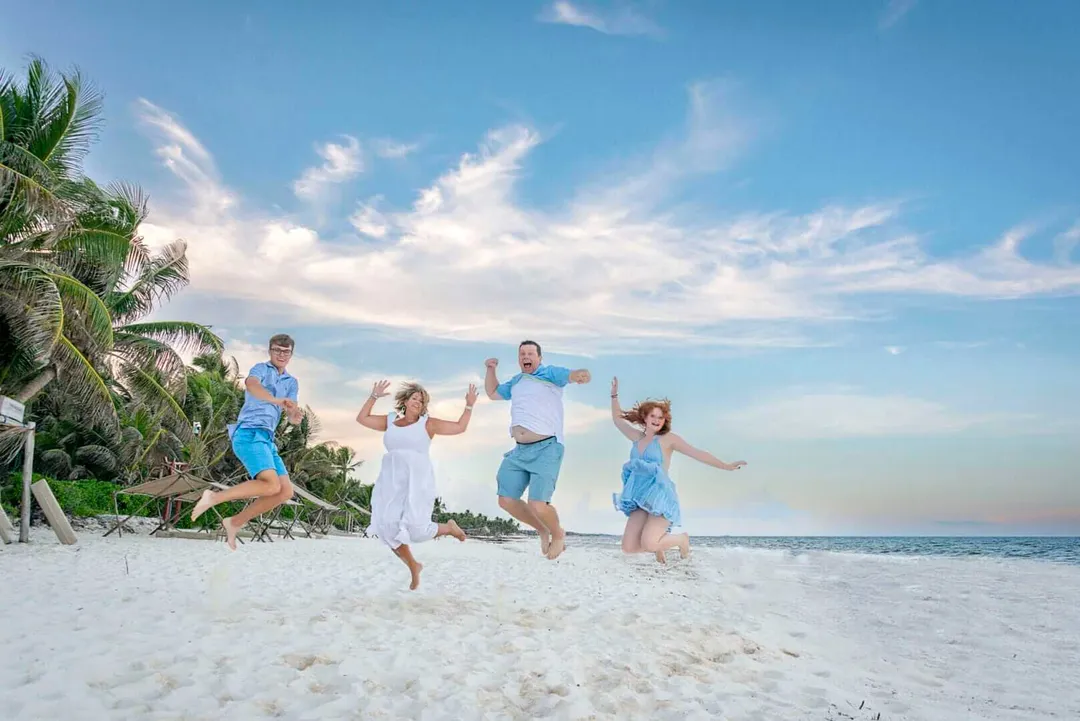 Family of five jumping on sandy beach with palm trees and ocean in background under partly cloudy sky