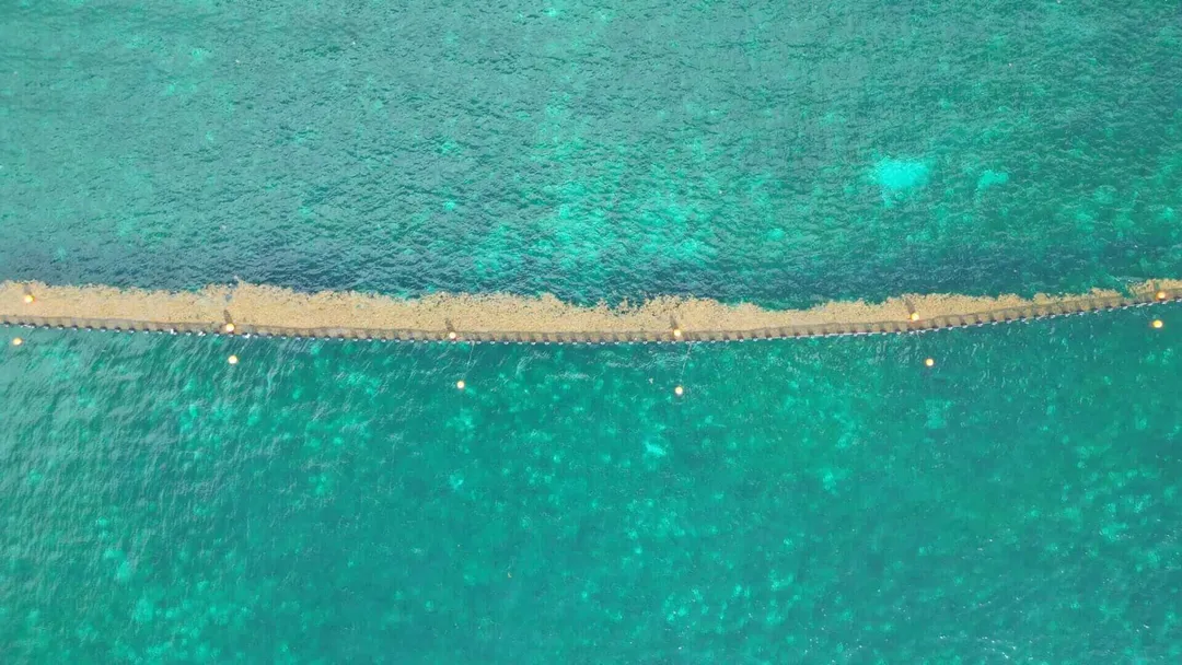 Aerial view of a narrow sandy barrier separating shallow turquoise water from deeper turquoise sea, with small boats and debris visible along the dividing line