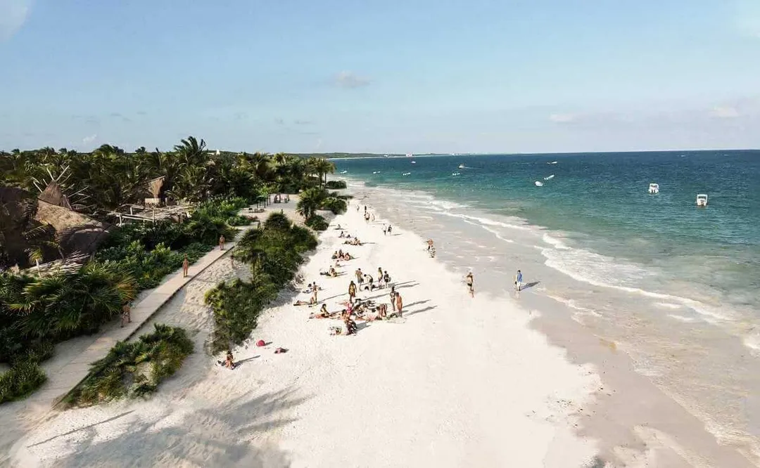 Beachgoers swim and relax on a sandy beach lined with tropical vegetation and structures along the shoreline