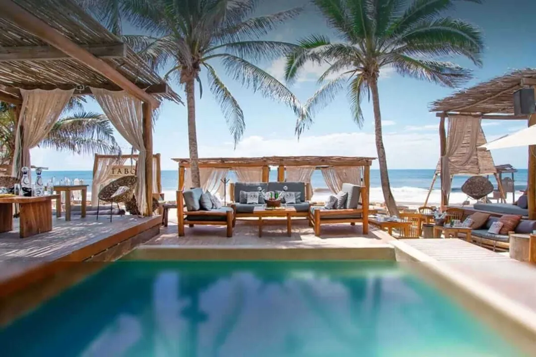 A beachfront resort lounge with a swimming pool, wooden cabanas with flowing curtains, palm trees, and ocean views under clear sky