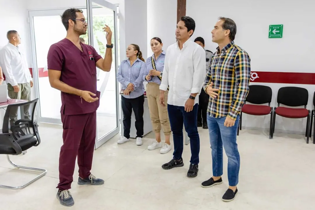 Man in burgundy scrubs gestures while speaking to a group of people listening in an indoor medical or clinic setting