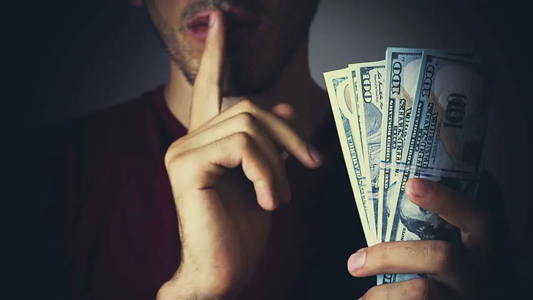 Person holding cash makes a shushing gesture with finger to lips against dark background