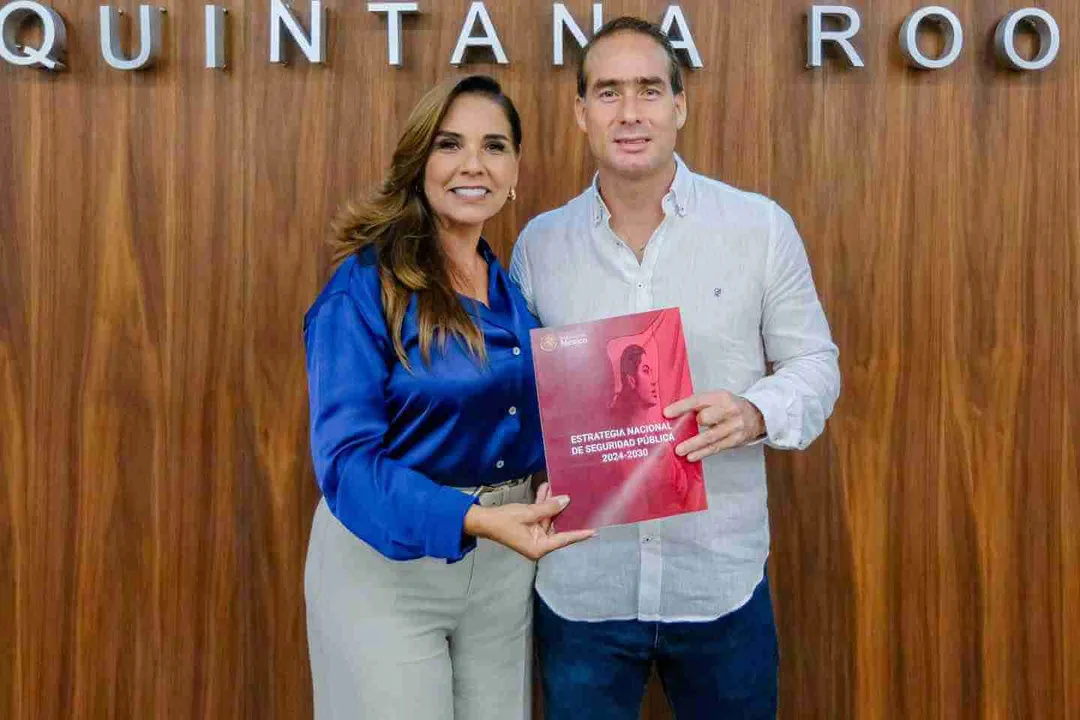 Tulum Mayor Joins National Security Strategy with Quintana Roo Leaders Two people stand together holding a red document in front of a wooden wall with partial text visible above them