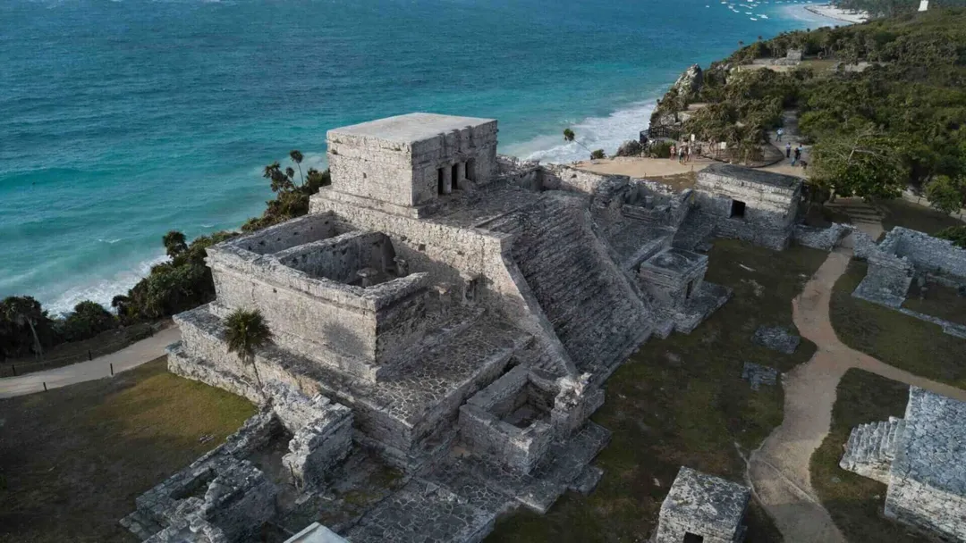 Tulum Faces Its Hardest Season in Decades Stone fortress ruins on a coastal cliff overlooking turquoise water and sandy beach, with vegetation and pathways visible around the structure