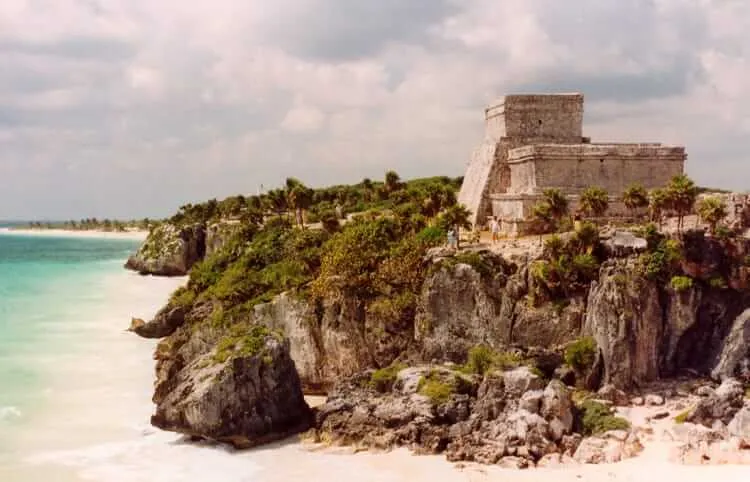 Stone fortress ruins sit atop a rocky cliff covered in vegetation above a turquoise beach and white sand