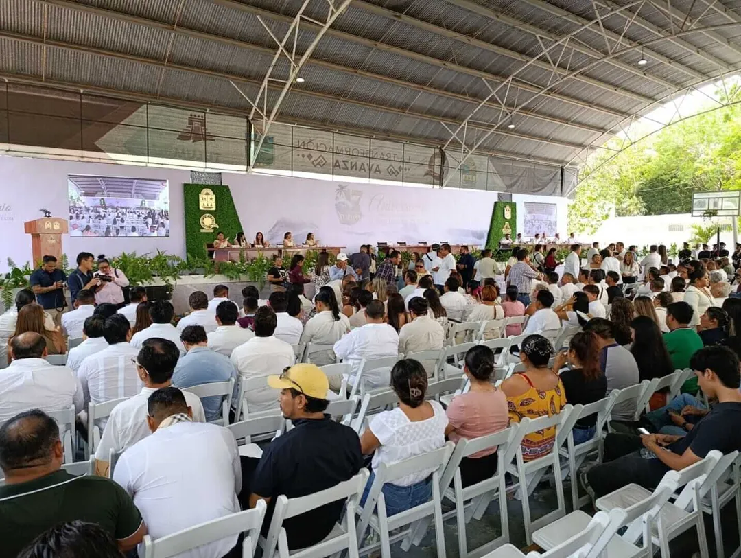 Building a Municipality That Honors Its Past Large crowd seated on white chairs in a covered outdoor venue with green decorative elements and a stage where speakers address the audience